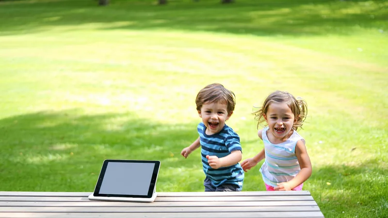 Children playing outside in summer sunshine, tablet resting unused on a garden bench